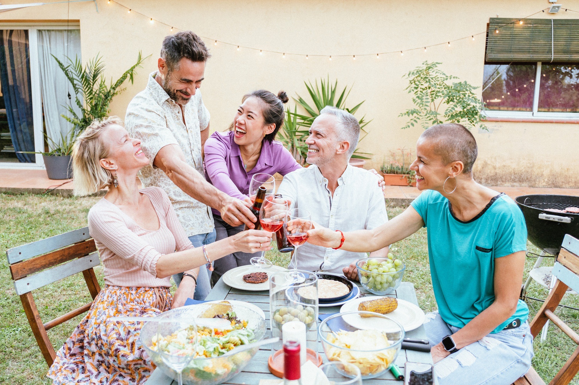 Group of happy middle-aged men and women toasting wine glasses at dinner event at house backyard.